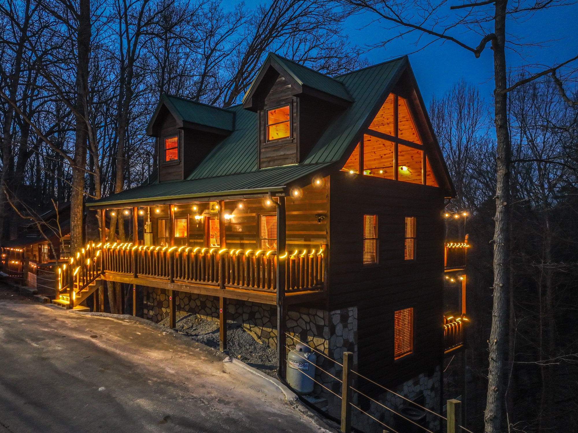 Aerial twilight view of the Bearadise cabin lit with string lights along its wraparound porch, surrounded by forest